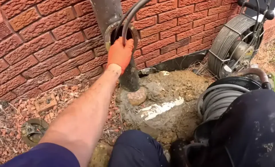 Gloved hand feeding a cable into a pipe access opening beside an inspection reel.