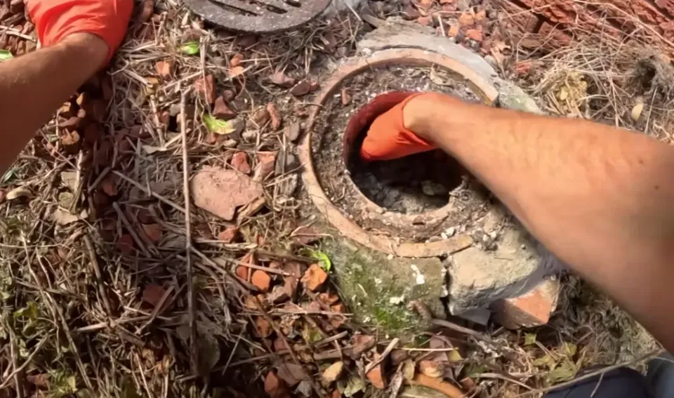 Gloved hand reaching into a round outdoor drain access opening.