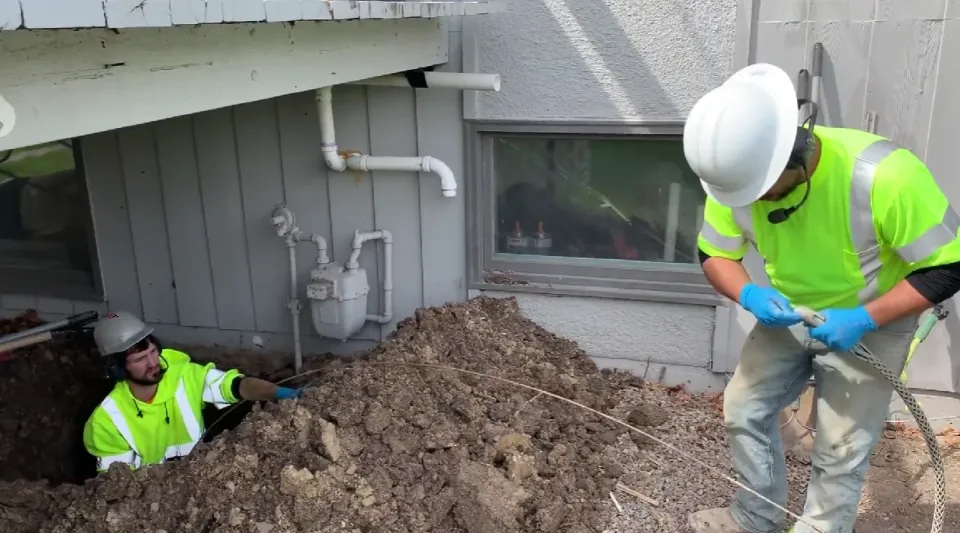 Workers handling a cable beside an excavation near a house.