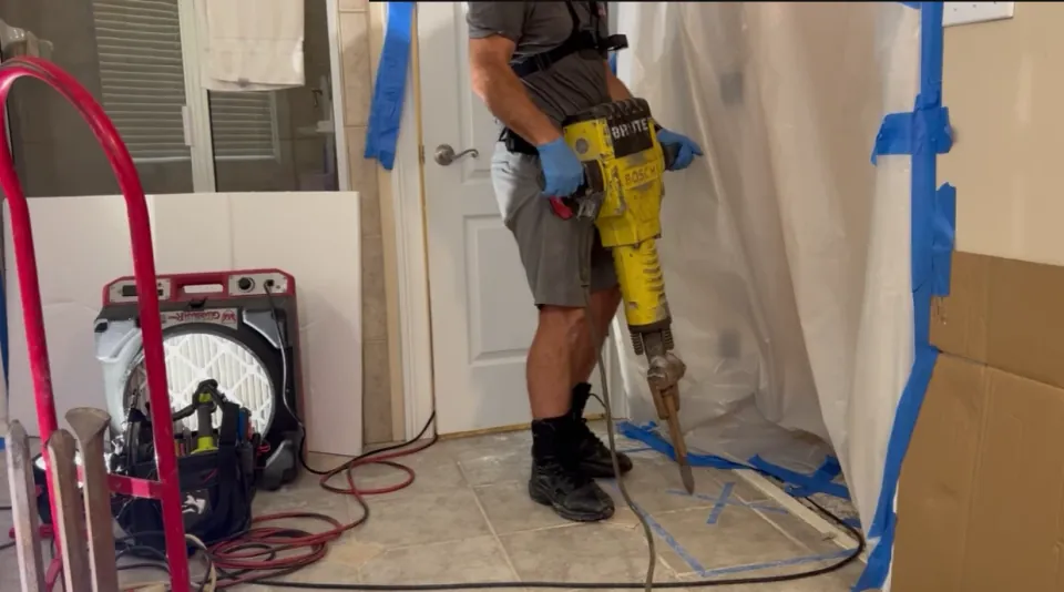 Worker using a demolition hammer on a marked section of tiled floor.